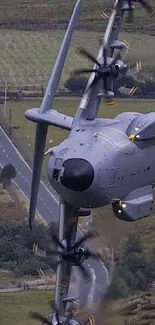 A gray aircraft flies over green fields with a road below.