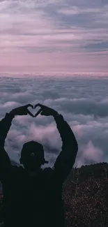Silhouette heart hands at sunset with clouds and mountains in the background.
