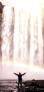 Man stands before a majestic waterfall with a vivid rainbow overhead.