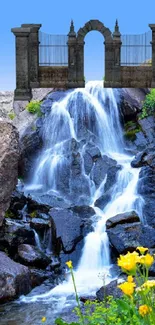 Waterfall beneath stone arch with wildflowers and blue sky.