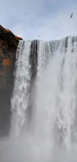 Majestic waterfall cascading down cliffs with a lone figure nearby, under a blue sky.