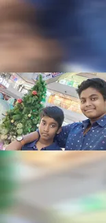 Two friends taking a selfie in a mall with a Christmas tree backdrop.