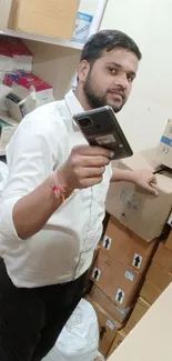 Man holds smartphone in box-filled room, wearing a white shirt.