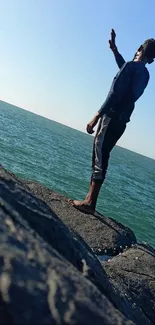 Person standing on a rocky ocean shore under a clear blue sky.