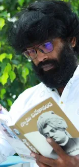 Bearded man reading a book against a leafy green backdrop.