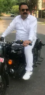 Man on a Royal Enfield motorcycle wearing white suit, riding on a peaceful road.