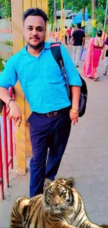 Man in blue shirt standing with tiger near pond in park.