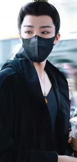 A masked person in black attire holding a coffee cup on the street.