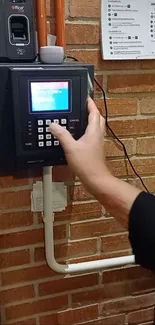 Hand interacting with a modern wall-mounted time clock on a brick wall.