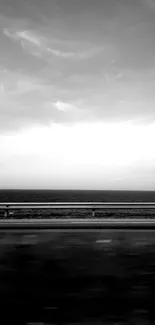 Monochrome view of road and sea under cloudy sky.