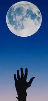 Silhouette hand reaching for the moon against a deep blue night sky.