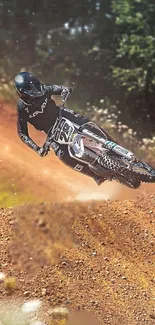 Motocross rider jumps dirt hill with scenic forest background.