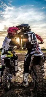 Two motocross riders share a moment under a sunset sky.