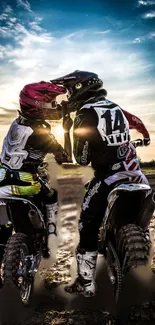 Two motocross riders sharing a moment under a sunset sky on a dirt track.