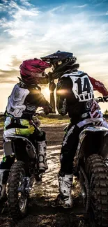 Two motocross riders in gear, sharing a moment against a sunset sky.