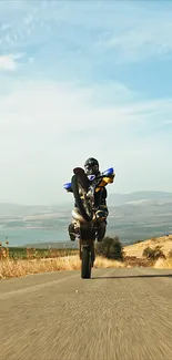 Motorcyclist performing a wheelie on a scenic road with clear blue sky.