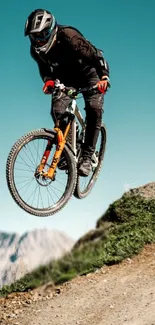 Biker jumps off a trail with mountain and blue sky backdrop.