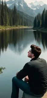 Person sitting by a tranquil mountain lake with forest and peaks.