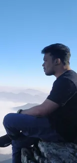 Person sitting on rocky cliff with blue sky above and misty mountains in the distance.