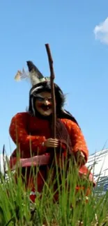 Witch figure on grassy field with snowy mountain and blue sky.