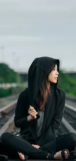 Hooded girl sitting on train tracks in urban setting.