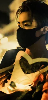 A masked figure holding a lit heart-shaped object at night.