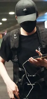 Masked person in black streetwear holding a phone with city lights in the background.