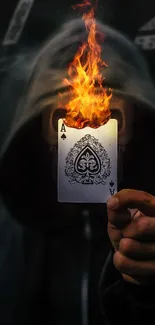 Hooded figure holding flaming playing card in dark setting.