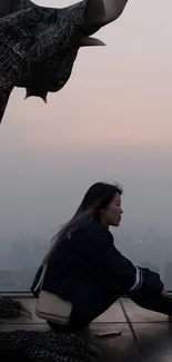 A person sits beside a dragon statue atop a rooftop with a sunset city view.