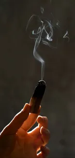 A hand delicately holds a burning stick emitting wispy smoke.