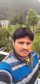 Man in a sweater taking a selfie with greenery and buildings in the background.