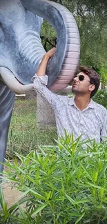 Man in sunglasses interacting with elephant statue amidst green foliage.