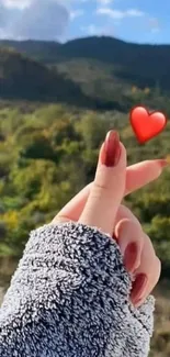 Hand gesture with heart over scenic green landscape.