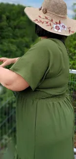 Person in an olive green outfit with a hat, enjoying a lush outdoor view.