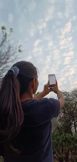 Person capturing nature with a smartphone against a cloudy sky.