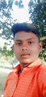 Young man in orange shirt taking a selfie outdoors with trees in the background.