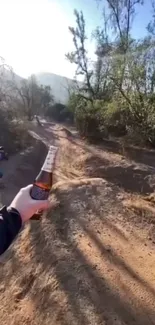 Hand holds drink bottle on forest trail under sunny sky.