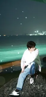 Person sits on a rock by the beach at night, with ocean waves and city lights.