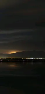 Nighttime view of mountains with city lights and reflection on a calm lake.