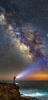 Person with flashlight under a starry sky by the ocean.