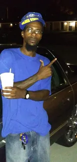 Man in blue shirt posing by car at night.