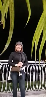 Person standing on a nighttime balcony with green leaves overhead.