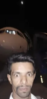 Nighttime selfie in front of a brightly lit airplane, set against a dark sky.