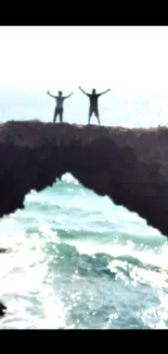 Two people stand on an ocean rock arch, against a blue sea background.