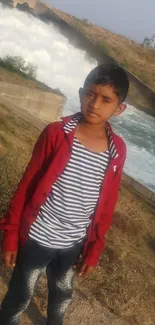 Boy in red jacket near a flowing river in a countryside setting.