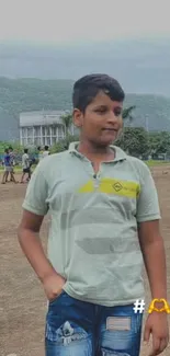 Boy standing in a natural outdoor setting with mountains and greenery.