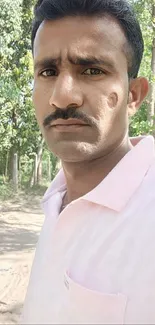 A man in a pink shirt outdoors with trees and greenery.