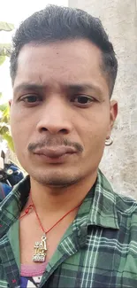 Man in green plaid shirt standing outdoors with motorbike and banana leaves.