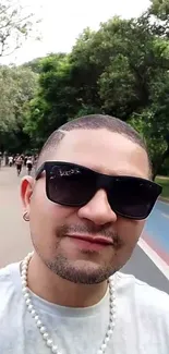 Person wearing sunglasses, taking a selfie in a green park.