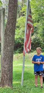 Child stands next to American flag in lush outdoor setting.